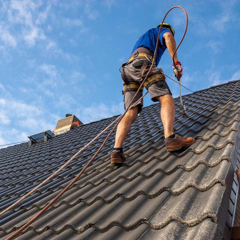 Couvreur : travaux en toute sécurité sur toit de tuiles Couvreur avec harnais et cordes de sécurité sur un toit de tuiles sombres, travaillant sous ciel bleu. Le mot 'safety' est visible sur sa chaussette.