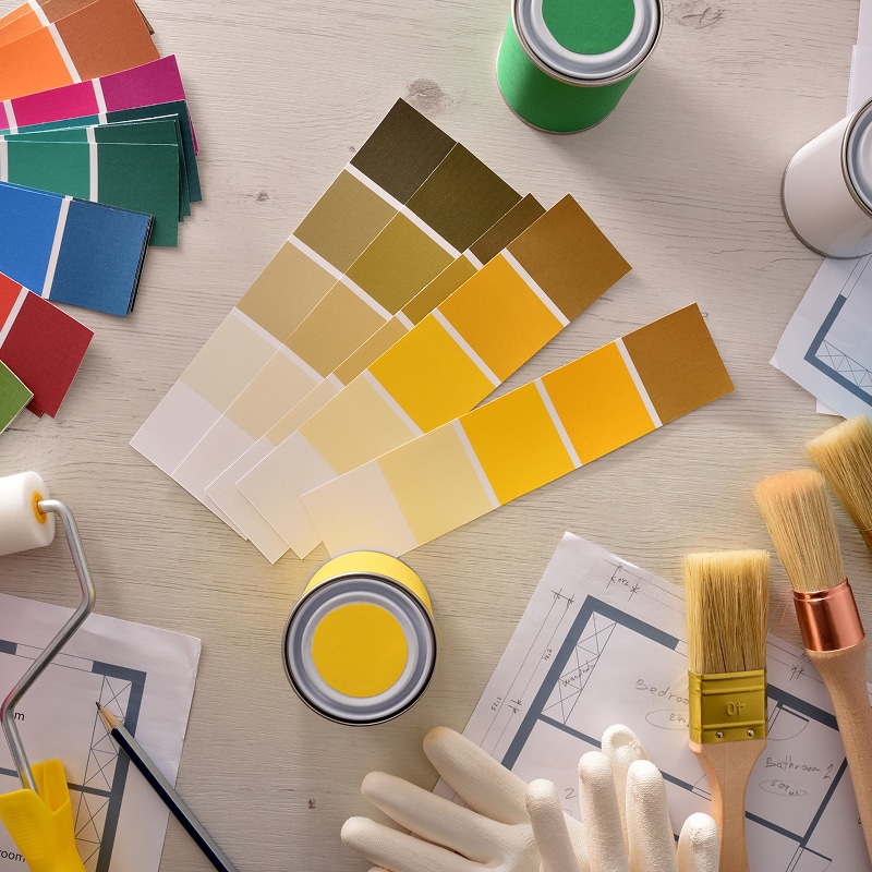 Peinture & Couleurs : Choisir sa déco intérieure facilement. Nuanciers de couleurs, pots de peinture jaune et vert, pinceaux, rouleau et plans de rénovation sur table bois clair. Préparation décoration intérieure.