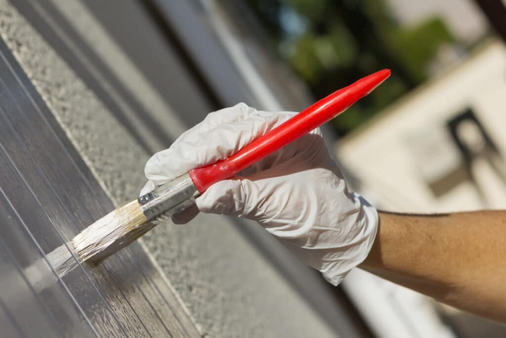 Peinture surface ondulée : Rénovation & protection extérieure Main gantée en train de peindre une surface ondulée translucide avec un pinceau à manche rouge pour rénovation et protection.