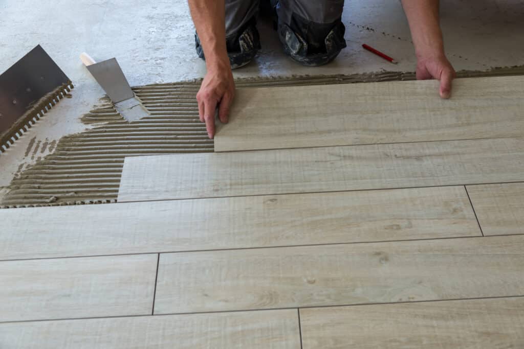 Pose de carrelage effet bois : Installation professionnelle Ouvrier agenouillé posant des carreaux de carrelage imitation bois clair sur une couche de colle au sol. Truelles visibles.