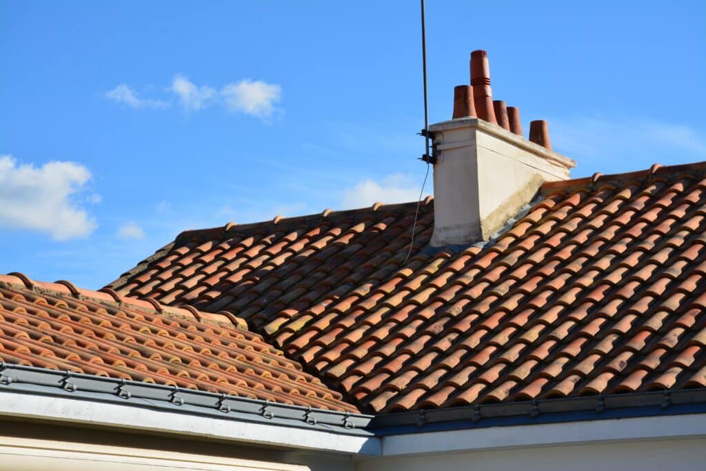 Toit en tuiles rouges avec cheminée et ciel bleu Toit en tuiles de terre cuite avec une cheminée et une antenne, sous un ciel bleu clair et des nuages blancs.