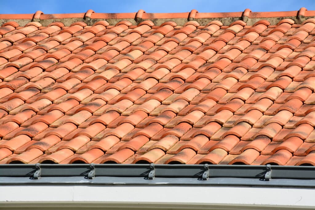 Toit en Tuiles de Terre Cuite Anciennes et Gouttière Grise Toit ancien en tuiles de terre cuite rouge-orangé sous un ciel bleu clair. Une gouttière grise est fixée à l'avant-toit blanc.